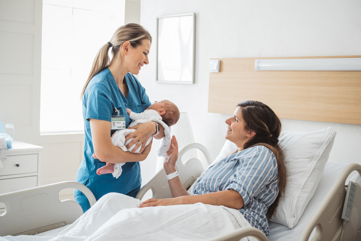 A labor and delivery nurse holding a newborn baby leans over to hand the baby to its mother.